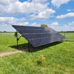 Panel słoneczny zamontowany na łące obok białego samochodu dostawczego z logo firmy instalującej, widok na oddalone pole i błękitne niebo z chmurami.