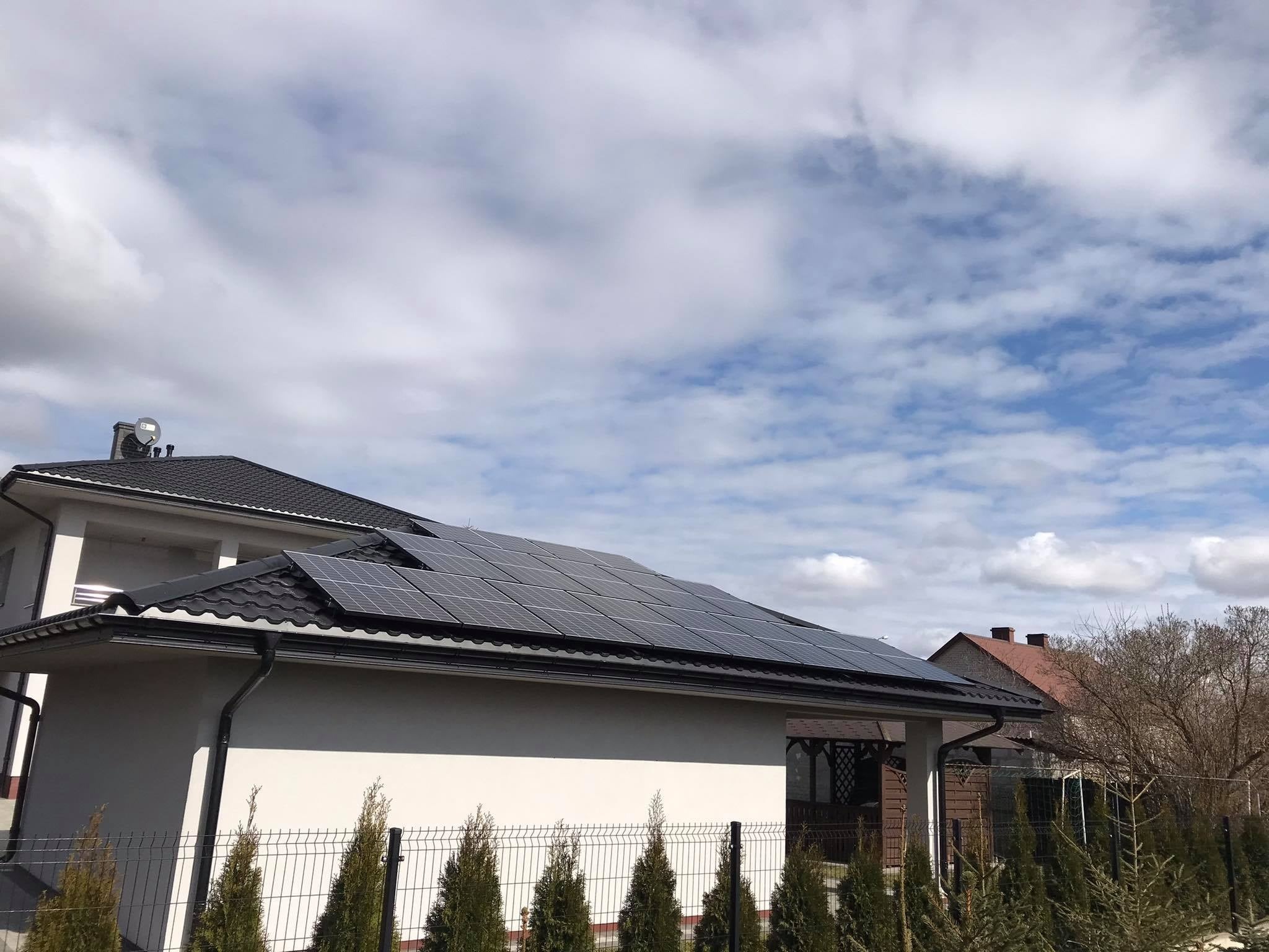 Panele na dachu domu jednorodzinnego z czarną dachówką, widok z poziomu ogrodzenia z tujami, błękitne niebo z chmurami w tle.