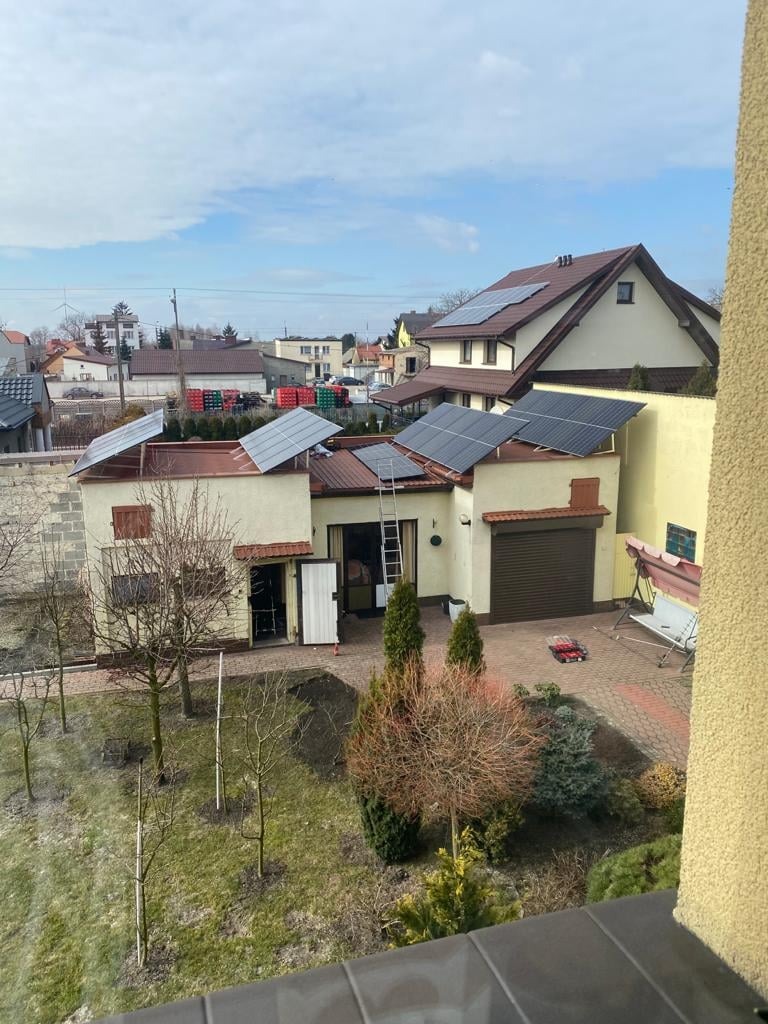 Widok z góry na dom z panelami słonecznymi na dachu, drabina oparta o budynek, ogród z młodymi drzewkami i kostką brukową.