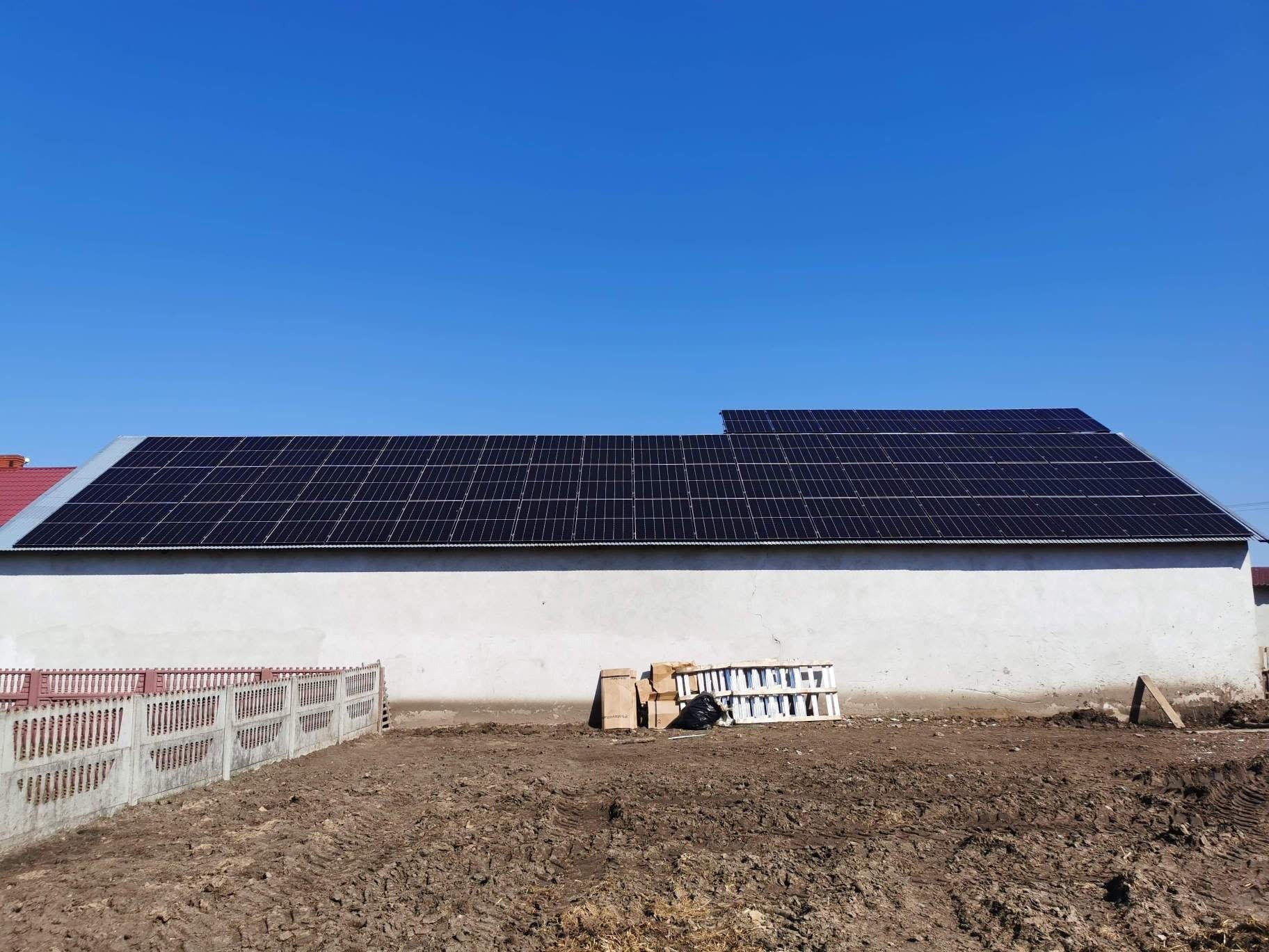 Instalacja fotowoltaiczna na dachu budynku gospodarczego z białymi ścianami, widoczna drabina oparta o ścianę i przygotowany teren.