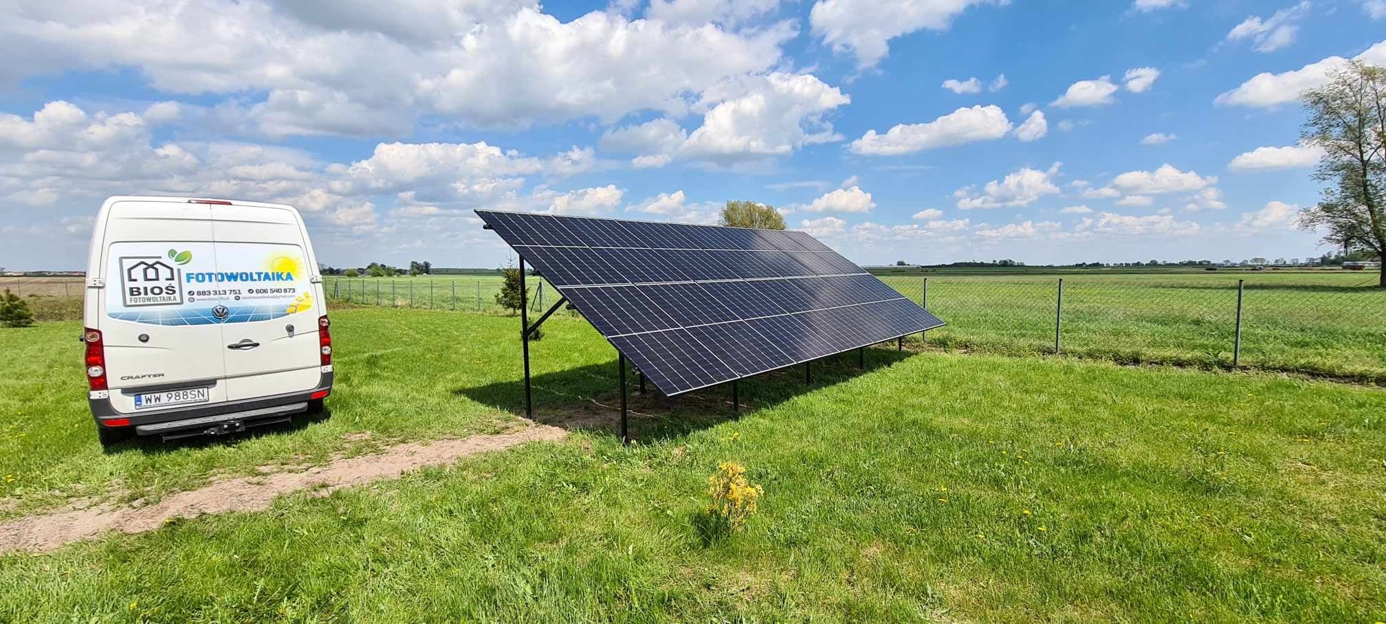 Panel słoneczny zamontowany na łące obok białego samochodu dostawczego z logo firmy instalującej, widok na oddalone pole i błękitne niebo z chmurami.