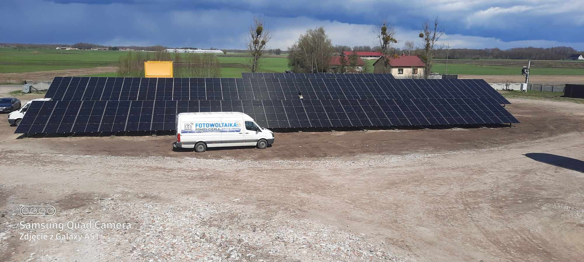 Rozległa instalacja paneli słonecznych na gruncie, z białym vanem firmowym z niebieskim logo i napisem, w tle zielone pola i burzowe chmury.