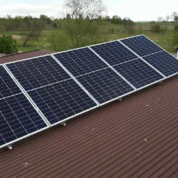 Rząd paneli fotowoltaicznych z białymi ramami zamontowany na brązowym, blaszanej dachu domu z widokiem na zielone pola w tle.