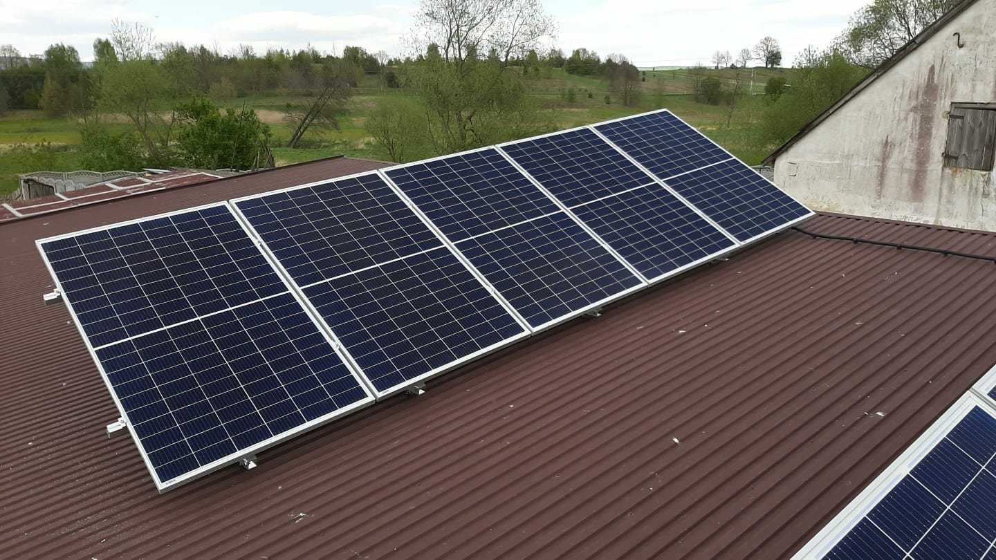 Rząd paneli fotowoltaicznych z białymi ramami zamontowany na brązowym, blaszanej dachu domu z widokiem na zielone pola w tle.