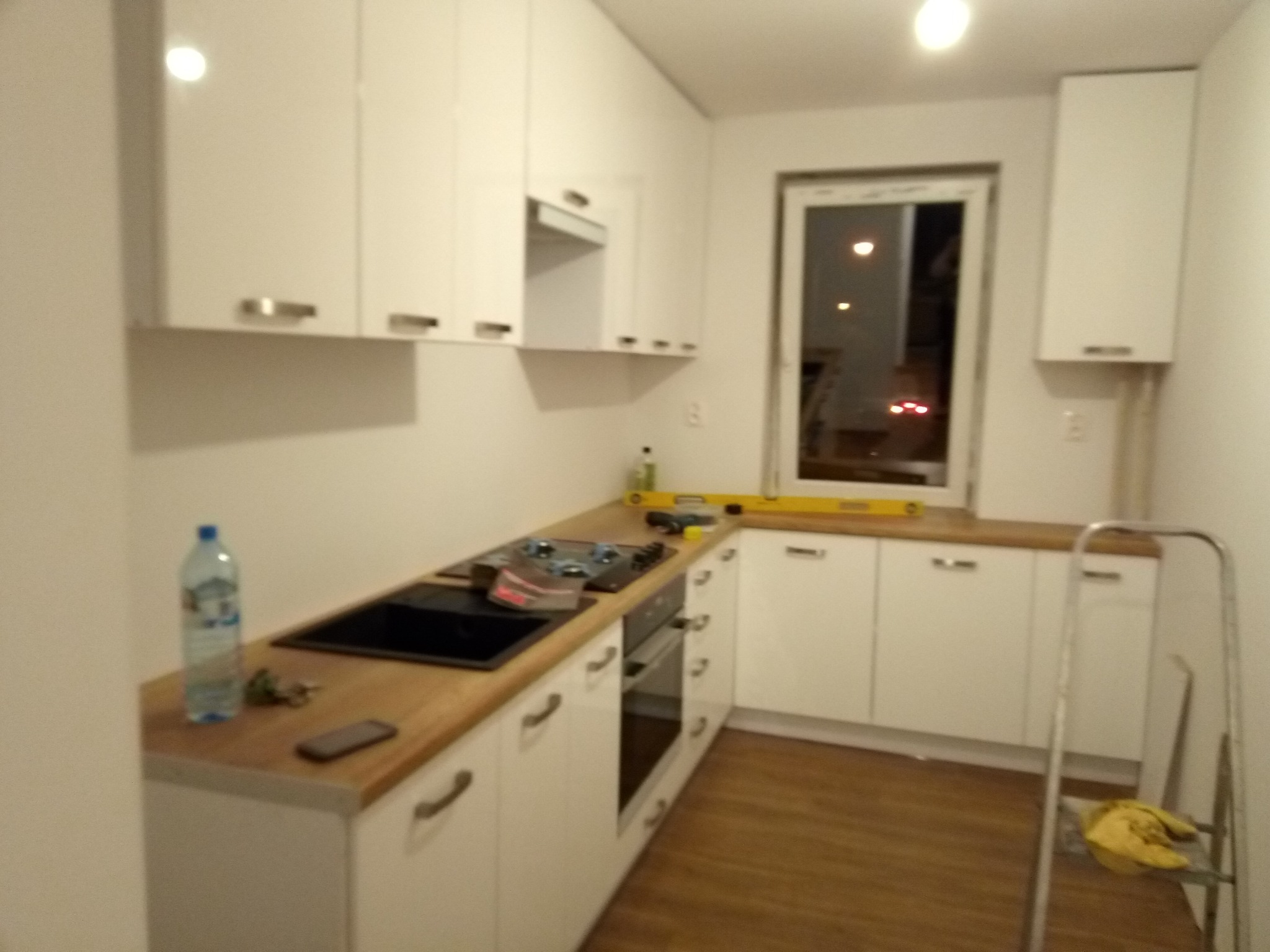 Montaż białej kuchni: widoczny fragment blatu z czarnym zlewem i płytą grzewczą, poziomica na blacie, butelka wody, drabina, wnętrze w trakcie wykańczania.