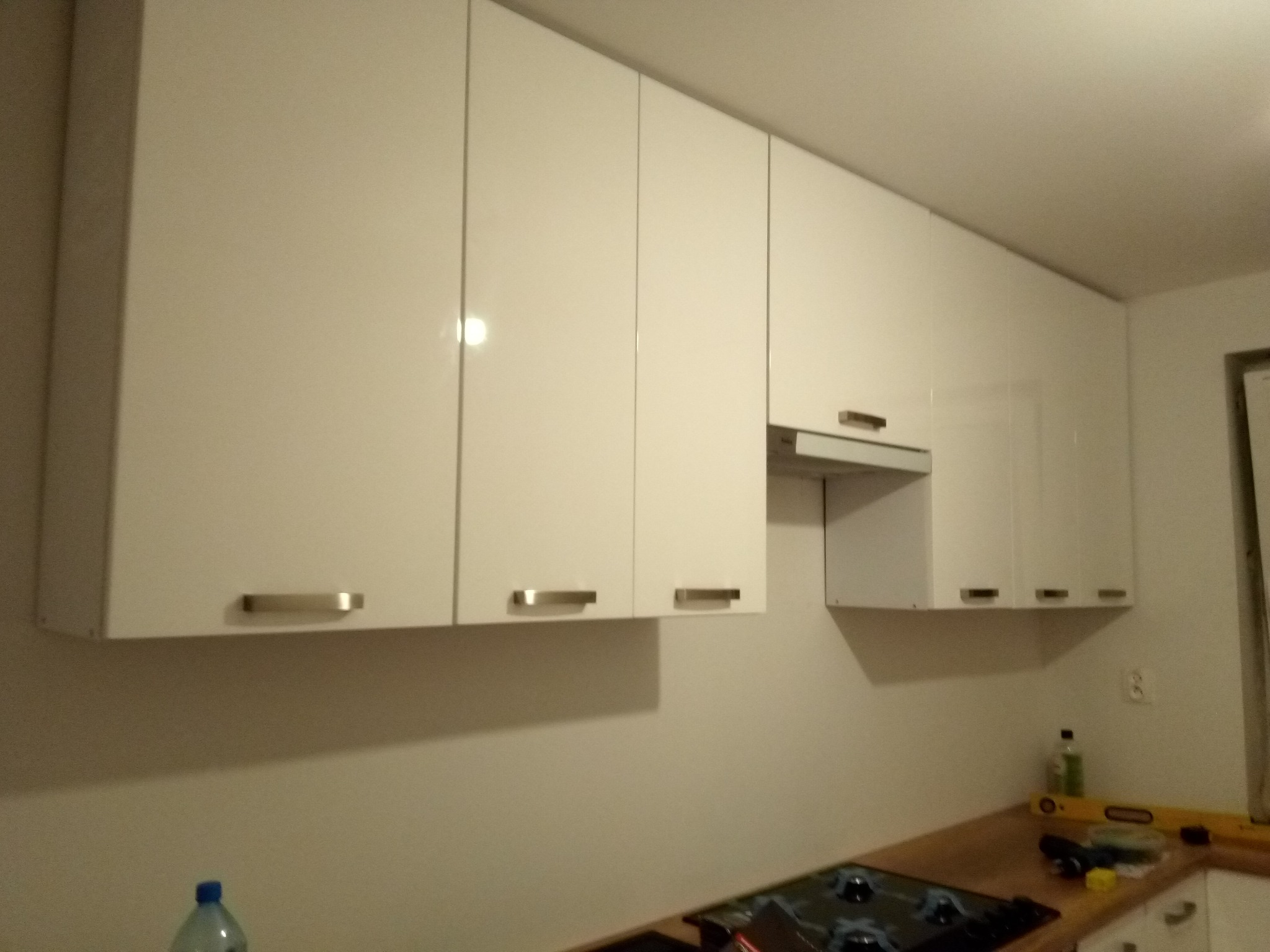 Białe, lakierowane szafki kuchenne wiszące z metalowymi uchwytami, nad kuchenką gazową i drewnianym blatem, widoczna butelka wody i poziomica.