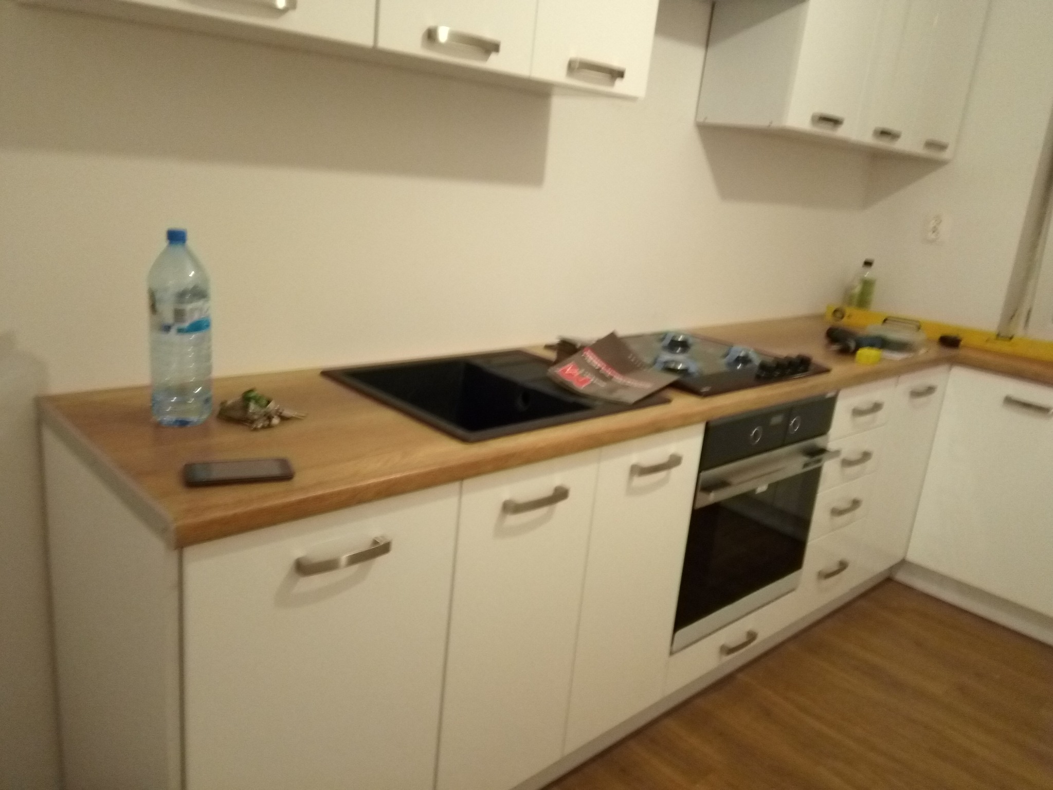 Fragment kuchni z białymi szafkami i drewnianym blatem, na którym leżą telefon, klucze, butelka wody, płyta indukcyjna i poziomica.
