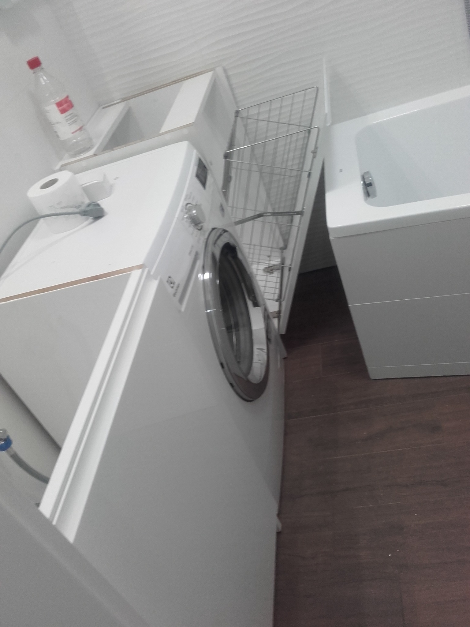 Biała pralka zabudowana pod blatem z wysuwanym koszem na pranie, obok wanna w minimalistycznej łazience z brązową podłogą.