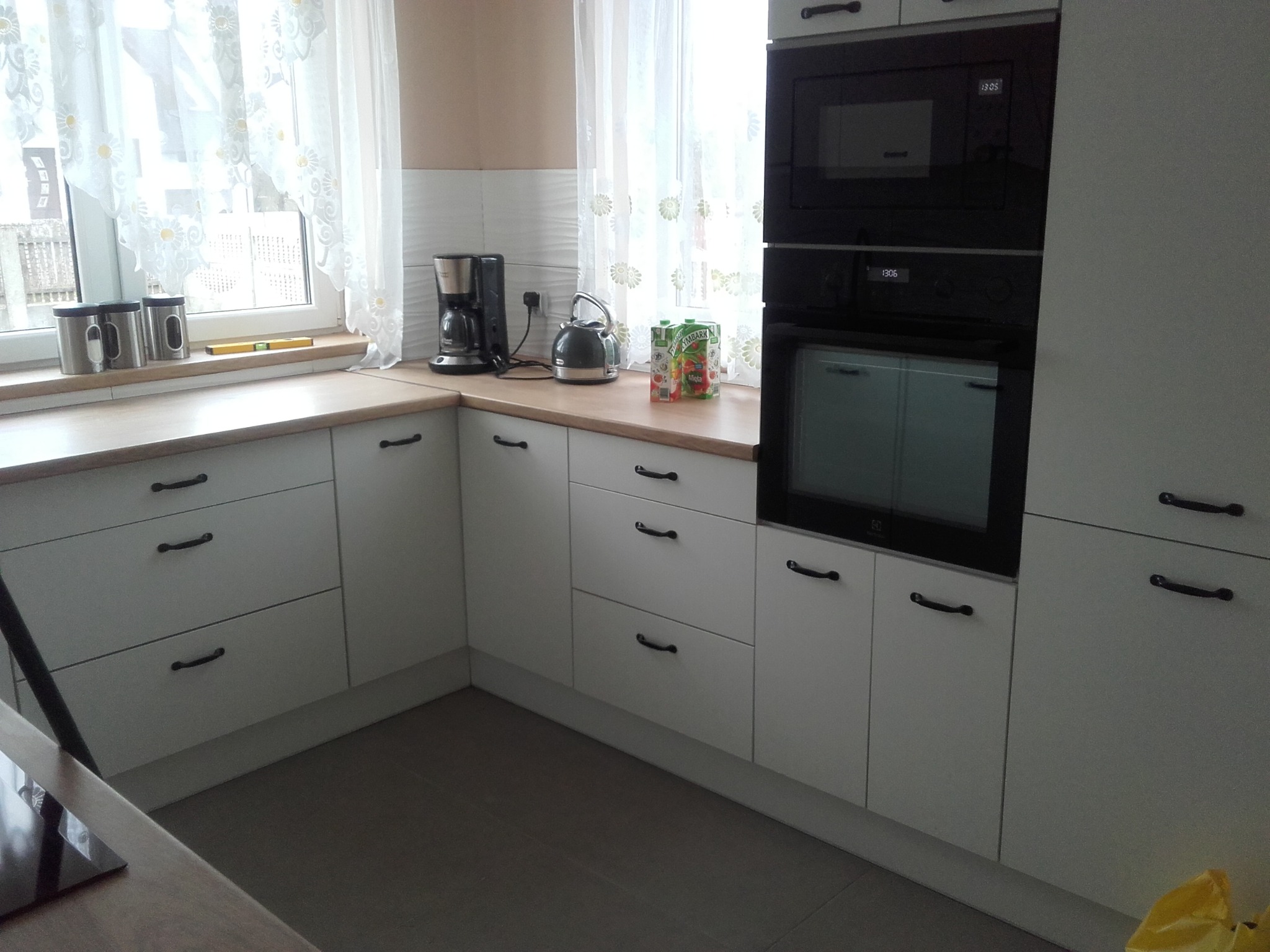 Jasna kuchnia z białymi frontami szafek i drewnianym blatem, czarne uchwyty, zabudowana kuchenka mikrofalowa i piekarnik, okno z firankami, czajnik elektryczny i ekspres do kawy na blacie.
