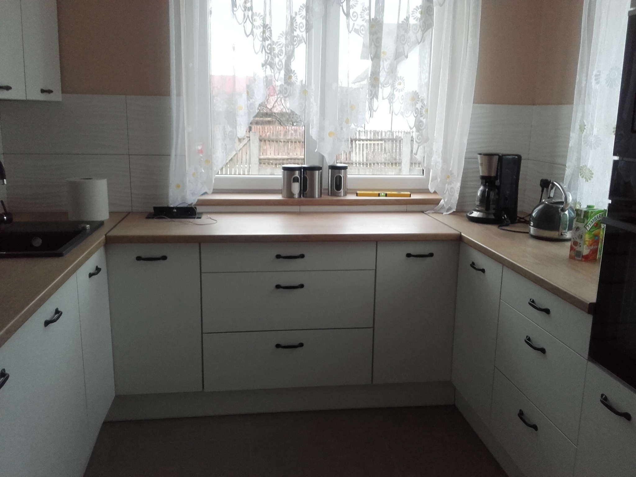 Jasna kuchnia z białymi szafkami i drewnianym blatem, widok na okno z firankami, na blacie czajnik, ekspres do kawy i pojemniki na przyprawy.