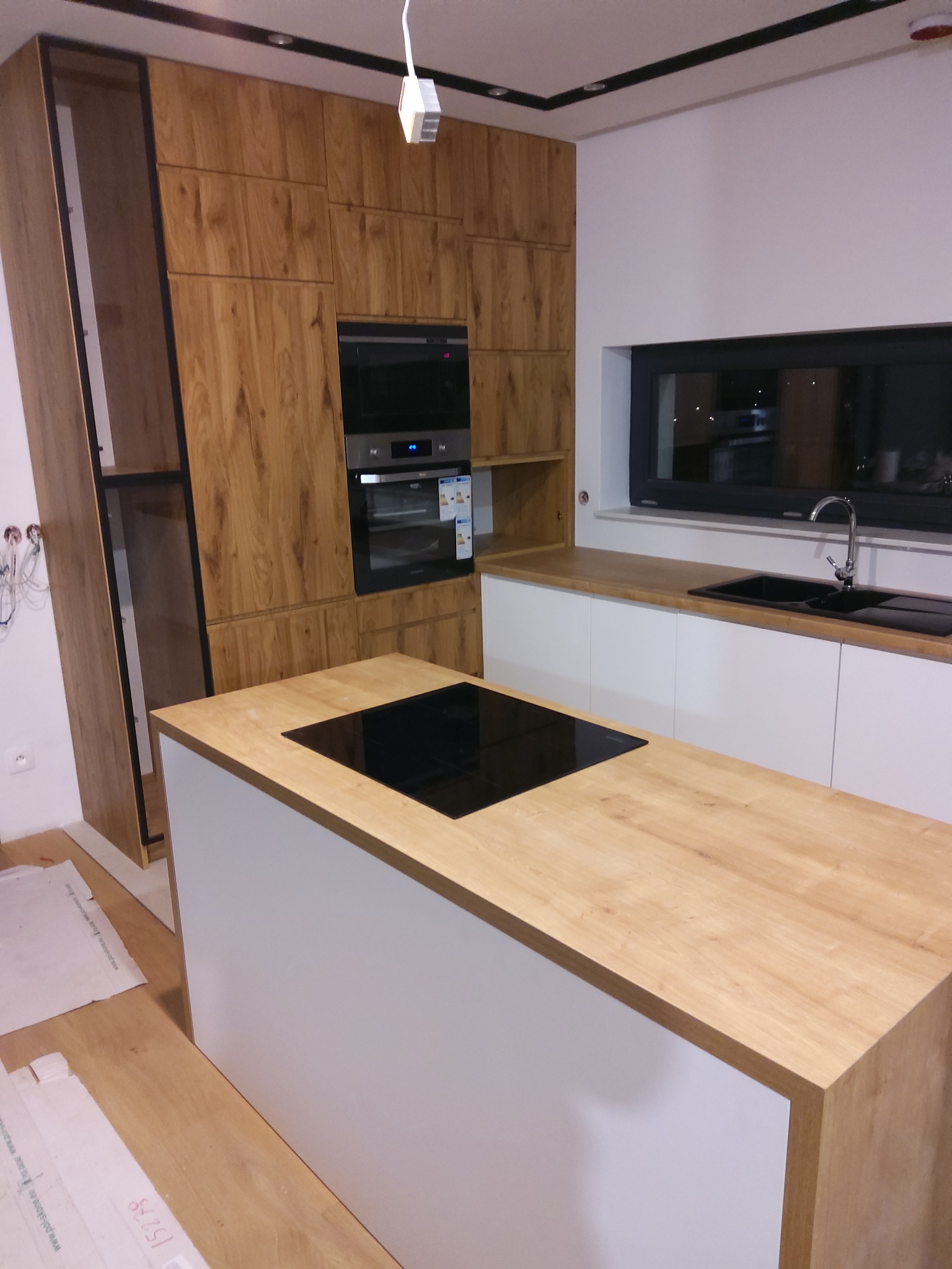 Nowoczesna kuchnia z drewnianymi frontami szafek i wyspą kuchenną z blatem imitującym drewno, płyta indukcyjna w wyspie, widoczne niedokończone prace wykończeniowe.