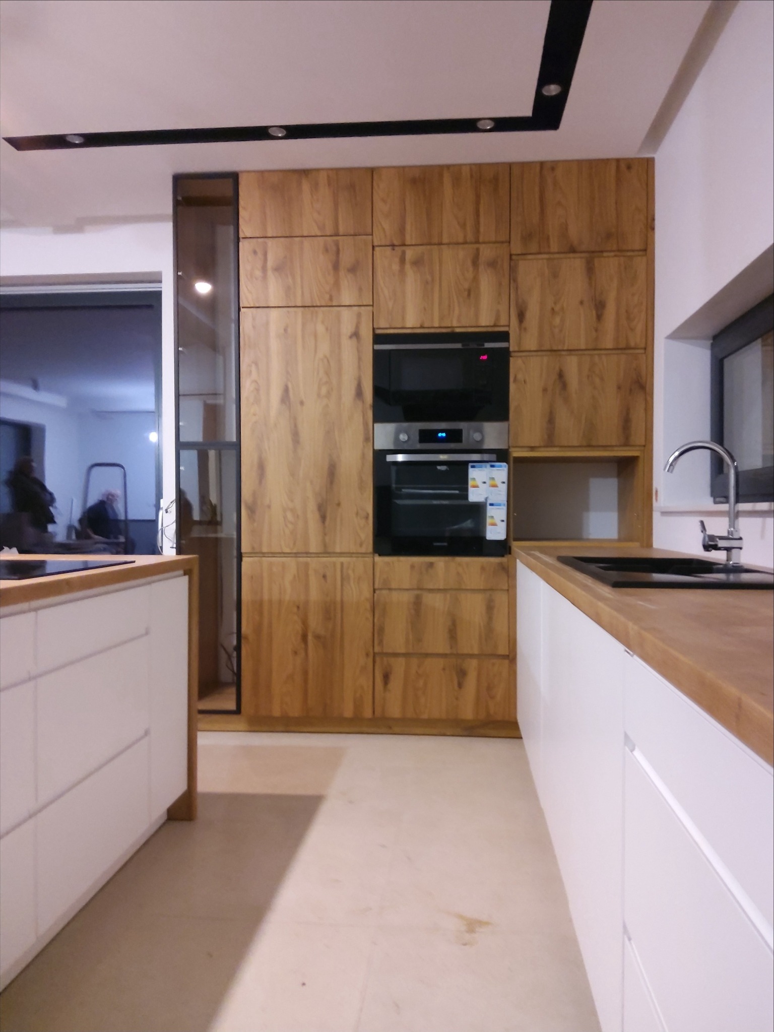 Nowoczesna kuchnia: zabudowa z drewnianymi frontami, białe szafki dolne z drewnianym blatem, piekarnik i mikrofalówka w zabudowie, widoczny zlew i bateria.