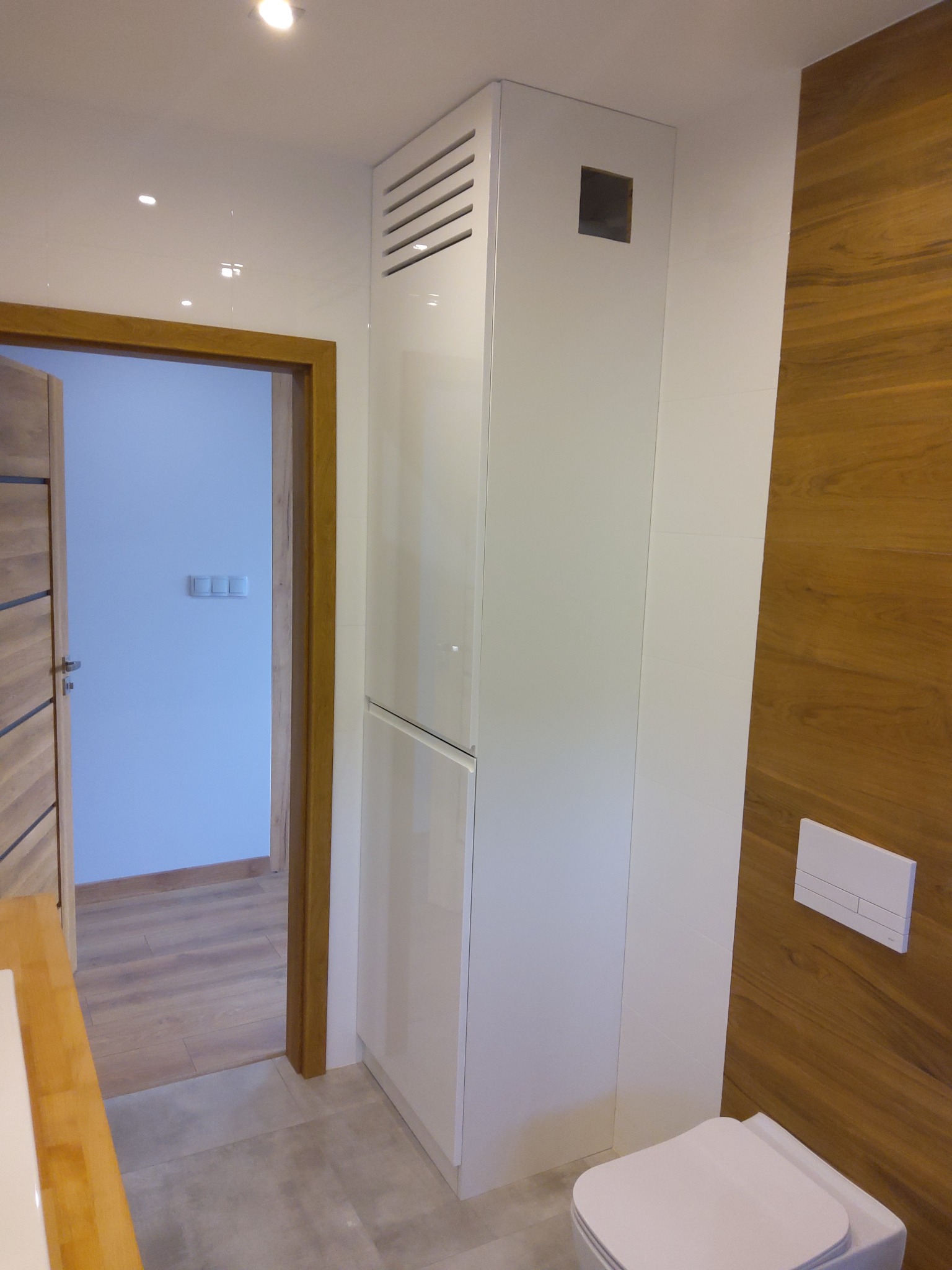 Wysoka, biała szafka łazienkowa z wentylacją w górnej części, zamontowana w Radomsku, obok drewnianej ściany i toalety podtynkowej.