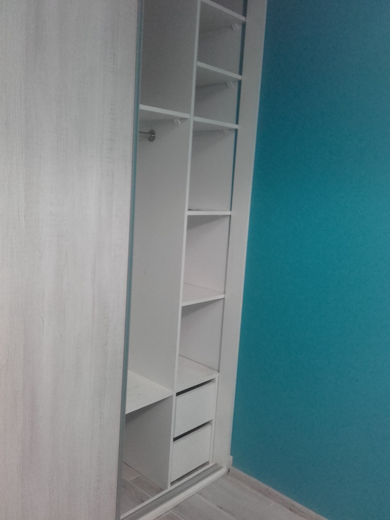 Wnętrze szafy wnękowej z jasnego drewna z półkami i szufladami, częściowo zasłonięte przesuwnymi drzwiami, na tle turkusowej ściany.