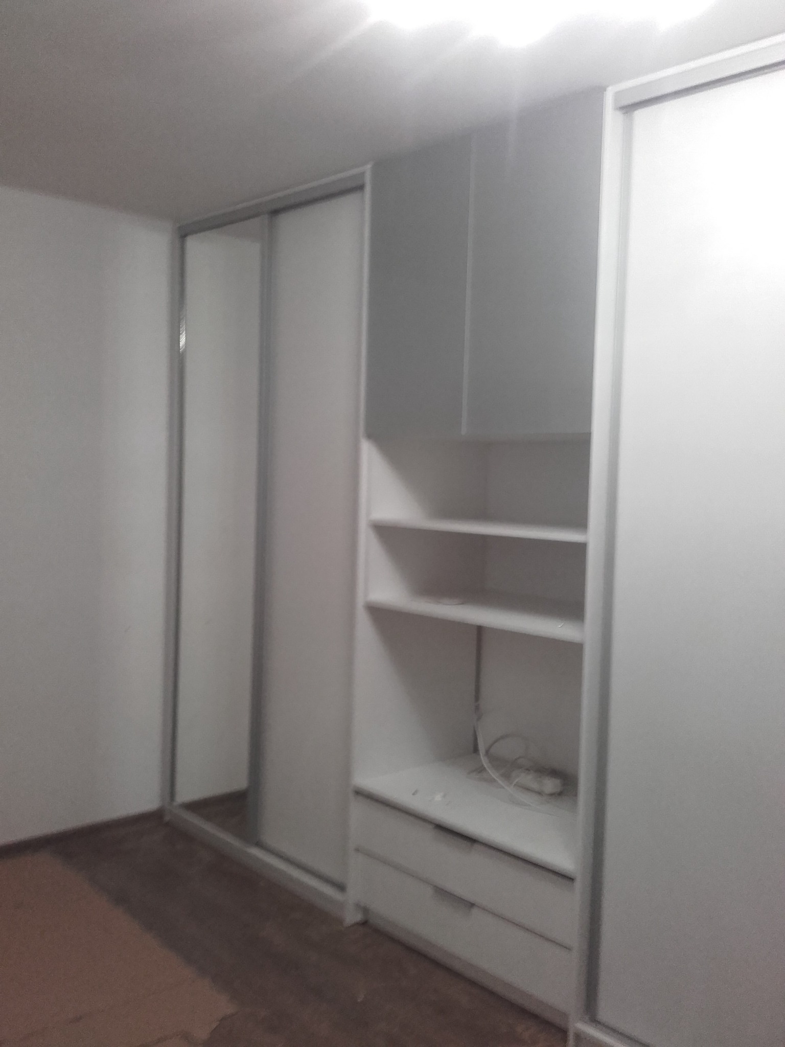 Biała szafa wnękowa z przesuwnymi drzwiami i otwartymi półkami, widoczne kable na jednej z półek, surowe wykończenie podłogi.