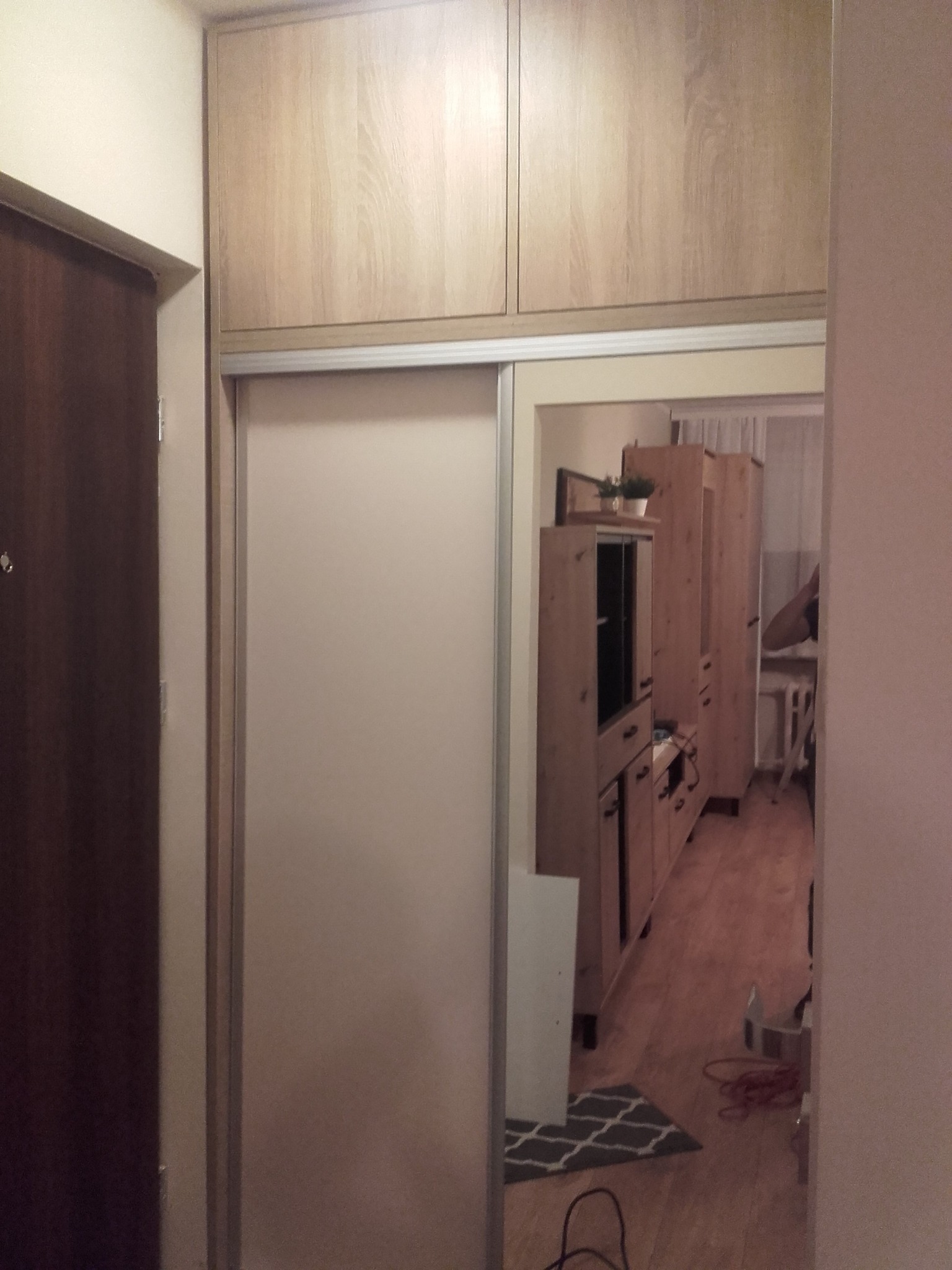 Wnętrze z zabudową: szafa przesuwna z górnymi szafkami w odcieniach drewna, widok przez otwarte drzwi na meble w salonie.