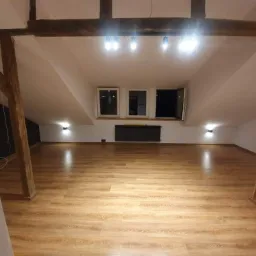 Poddasze po remoncie z drewnianymi belkami konstrukcyjnymi, panelami podłogowymi w odcieniach brązu i beżu, białymi ścianami oraz oknami dachowymi z widocznym grzejnikiem pod nimi.