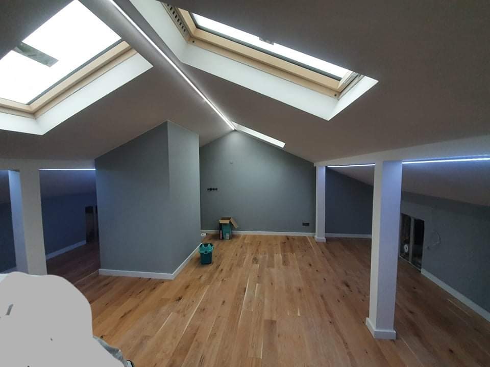 Przestronne poddasze po remoncie z drewnianą podłogą, szarymi ścianami, oknami dachowymi i subtelnym oświetleniem LED wzdłuż krawędzi sufitu.