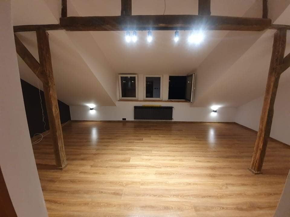 Poddasze po remoncie z drewnianymi belkami konstrukcyjnymi, panelami podłogowymi w odcieniach brązu i beżu, białymi ścianami oraz oknami dachowymi z widocznym grzejnikiem pod nimi.