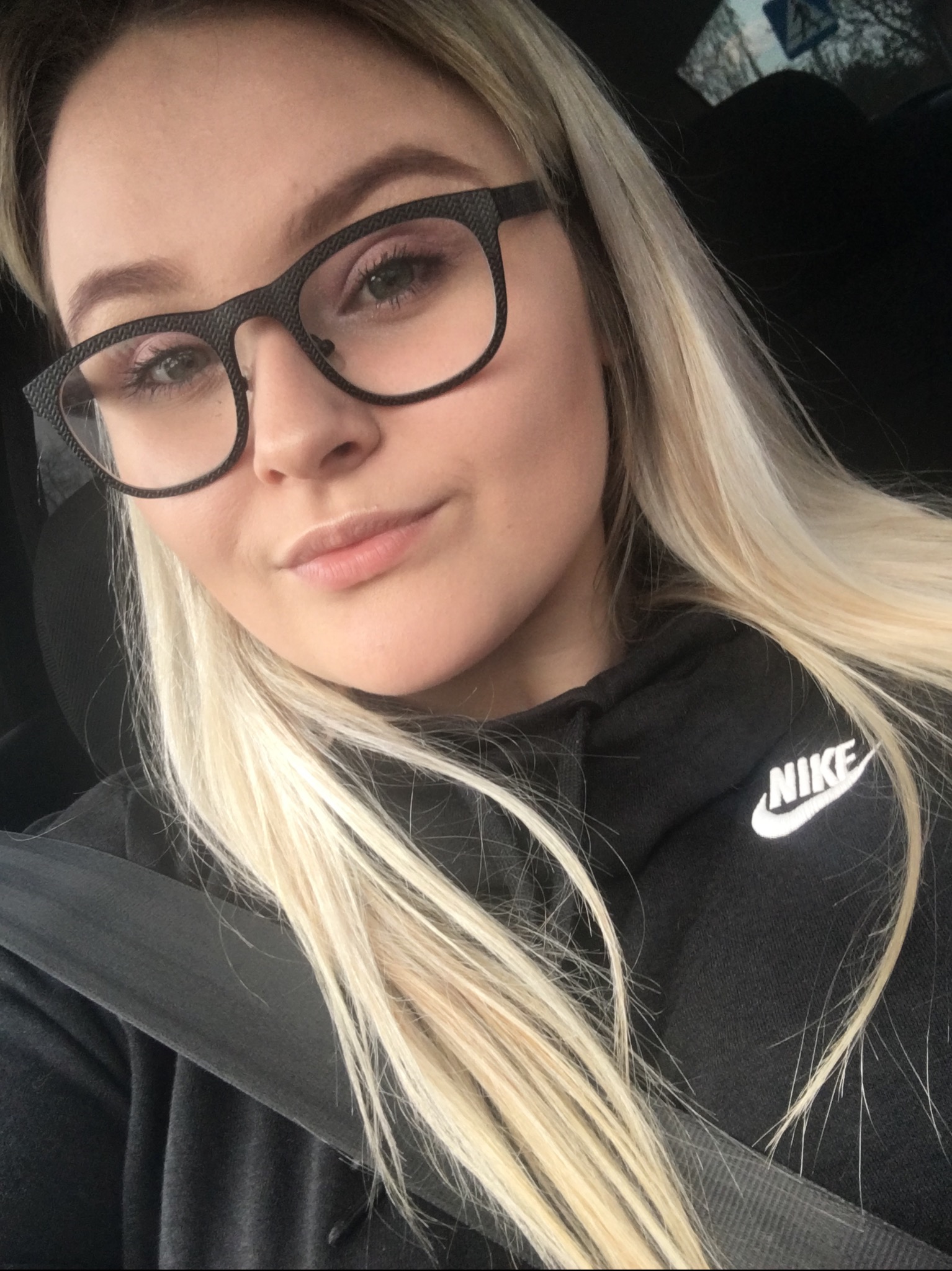 Blondwłosa kobieta w czarnych okularach o geometrycznej fakturze i czarnej bluzie Nike, zapięta pasem bezpieczeństwa, robi selfie.