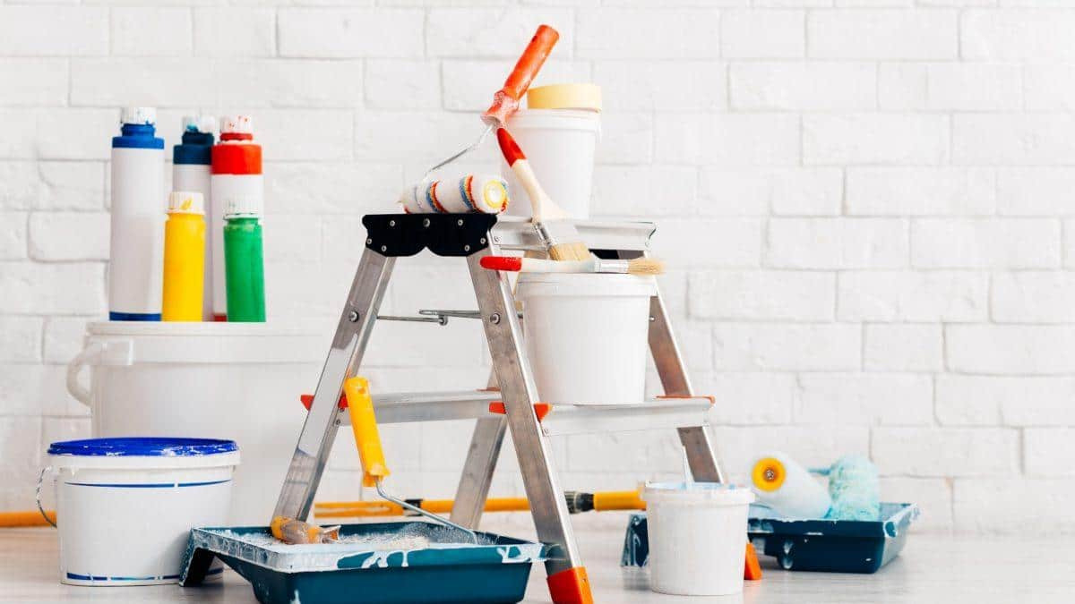 Metalowa drabina z farbą, wałkami, pędzlami i kuwetami na tle białej ściany z cegieł.