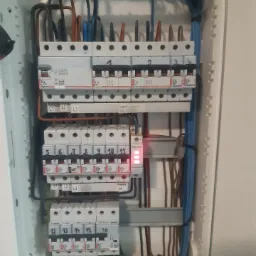 Otwarta skrzynka rozdzielcza z zainstalowanymi bezpiecznikami i przewodami o różnych kolorach, niektóre z numerami od 1 do 16, z widocznymi elementami elektronicznymi i złączkami.