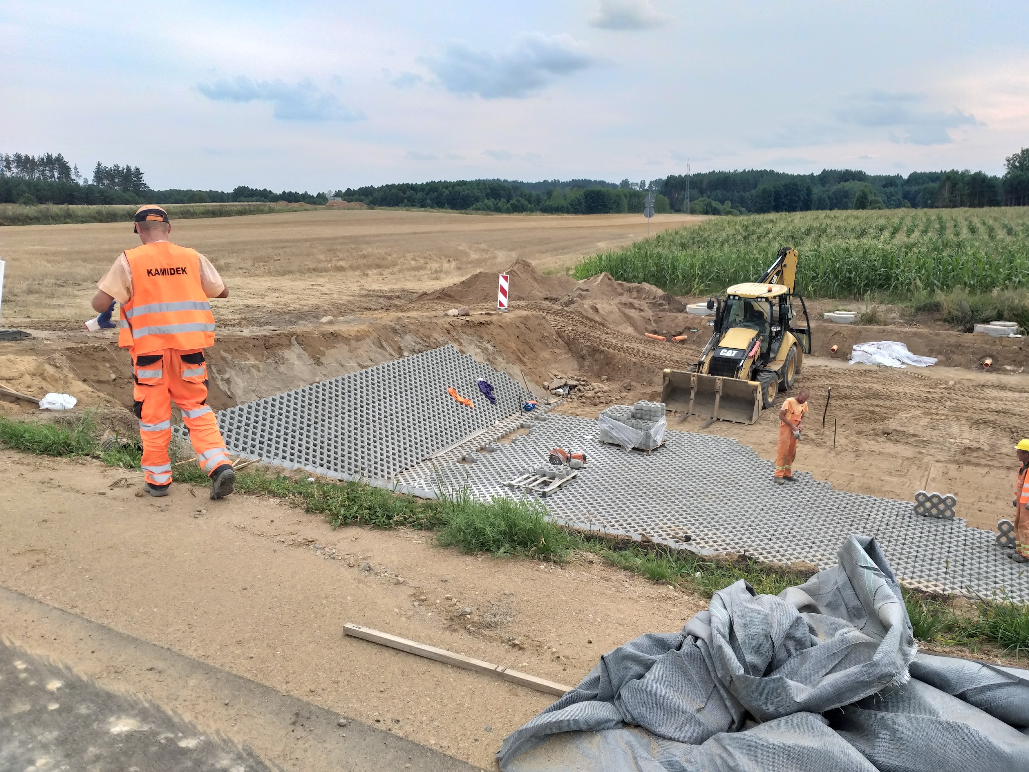Prace ziemne z wykorzystaniem ażurowych płyt betonowych na tle pola kukurydzy i koparki, widoczni pracownicy w pomarańczowych kombinezonach.