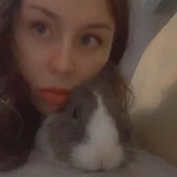 Kobieta przytula szarego królika z białą plamą na pysku, leżąc na jasnej pościeli.