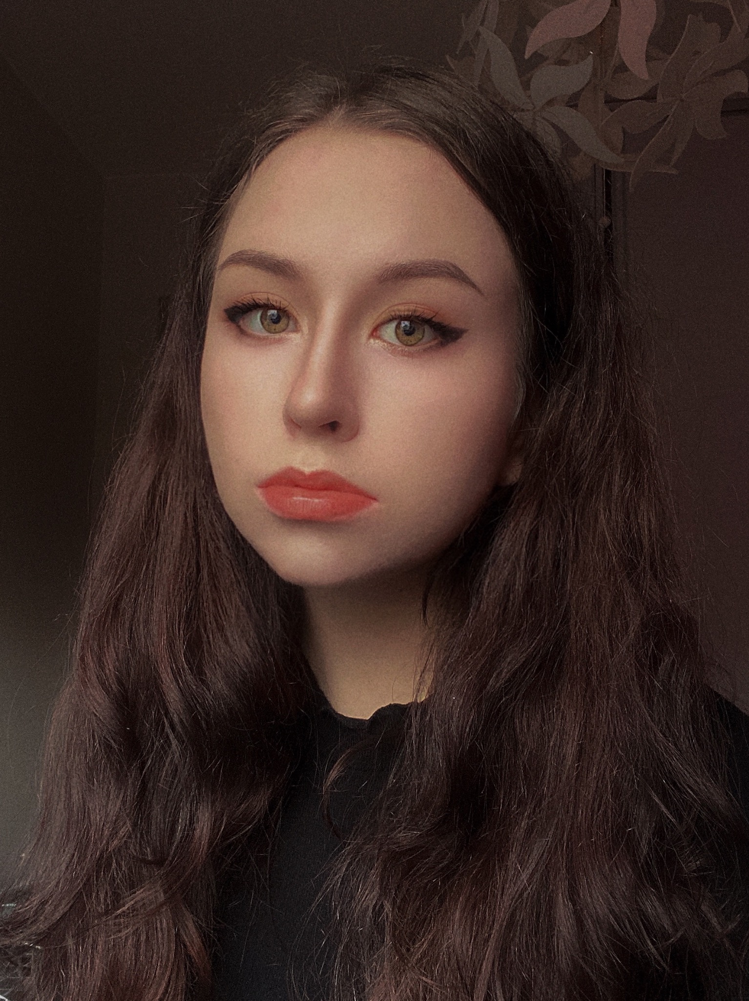 Portret młodej kobiety z długimi, brązowymi włosami, subtelnym makijażem oczu z eyelinerem i pomarańczową szminką, na ciemnym tle z delikatnym wzorem kwiatowym.