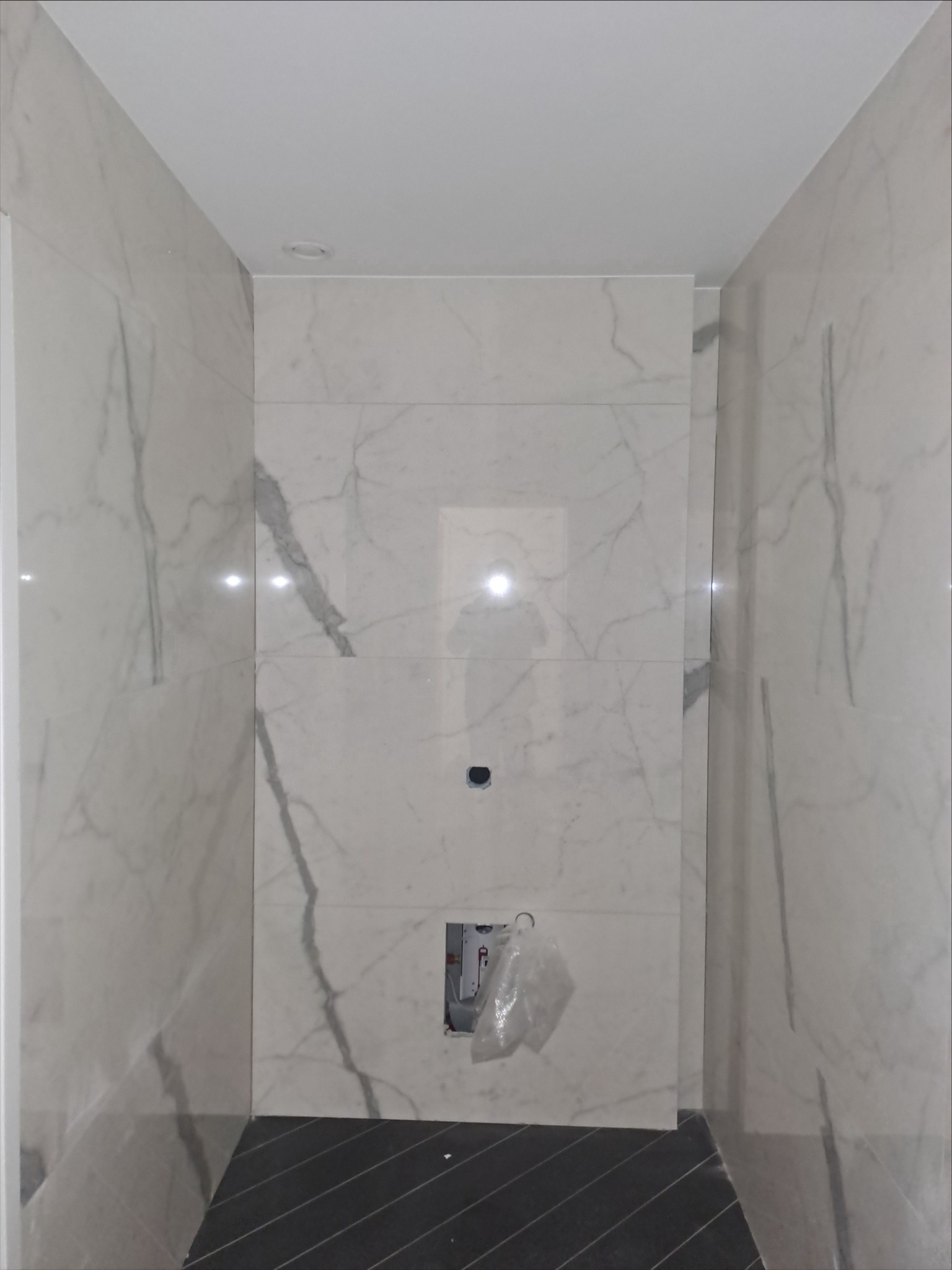 Wnętrze łazienki w trakcie wykańczania: marmurowe płytki ścienne z wyraźnym użyleniem, ciemna podłoga, widoczne instalacje. Minimalistyczny design w trakcie realizacji.