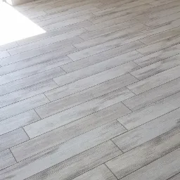 Ułożona pod kątem podłoga z płytek ceramicznych imitujących drewno o jasnoszarym odcieniu, z widoczną fugą i fragmentem ściany z przypodłogową listwą ceramiczną, oświetlona naturalnym światłem...