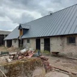 Budynek gospodarczy z szarego kamienia z nowym, szarym dachem blaszanym, widoczne fragmenty gruzu i materiałów budowlanych przed budynkiem, pochmurne niebo w tle.