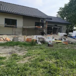 Dwuosobowa ekipa na rusztowaniu, tynkowanie elewacji domu jednorodzinnego z ciemną dachówką, widoczne materiały budowlane i narzędzia na trawniku.