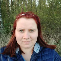 Portret kobiety z rudymi włosami i zielonymi oczami na tle lasu i krzewów w świetle popołudniowego słońca.