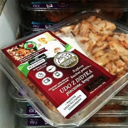 Opakowanie krojonego, pieczonego uda z indyka w przezroczystym plastikowym pojemniku z etykietą Wielkopolski Indyk, widoczne inne opakowania w tle.