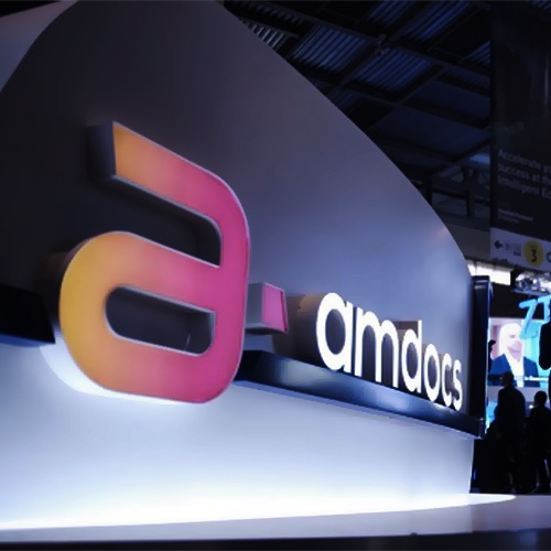 Podświetlane, przestrzenne litery tworzące logo firmy 'amdocs' na ciemnoszarym tle, z widocznymi sylwetkami ludzi w tle.