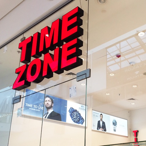 Czerwony, podświetlany szyld 'TIME ZONE' z literami przestrzennymi, zamontowany nad witryną sklepu w galerii handlowej. W tle widoczne reklamy zegarków.