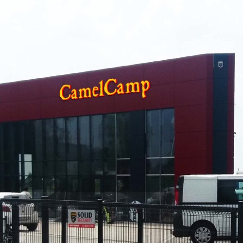 Budynek z podświetlanym szyldem 'CamelCamp' wykonanym z liter przestrzennych, widoczny zza ogrodzenia.