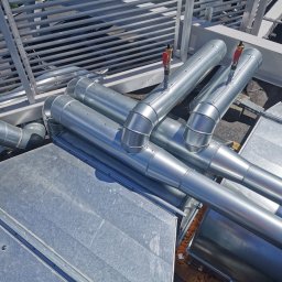 MDM Damian Grabiwski - Błyszczące, metalowe rury wentylacyjne z zaworami na dachu, widziane z góry, z elementami konstrukcji wsporczej w tle.