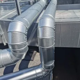 Dwa równoległe, ocynkowane przewody wentylacyjne z licznymi łączeniami nitowanymi, zamontowane na dachu pokrytym papą, widoczne elementy konstrukcji wsporczej.