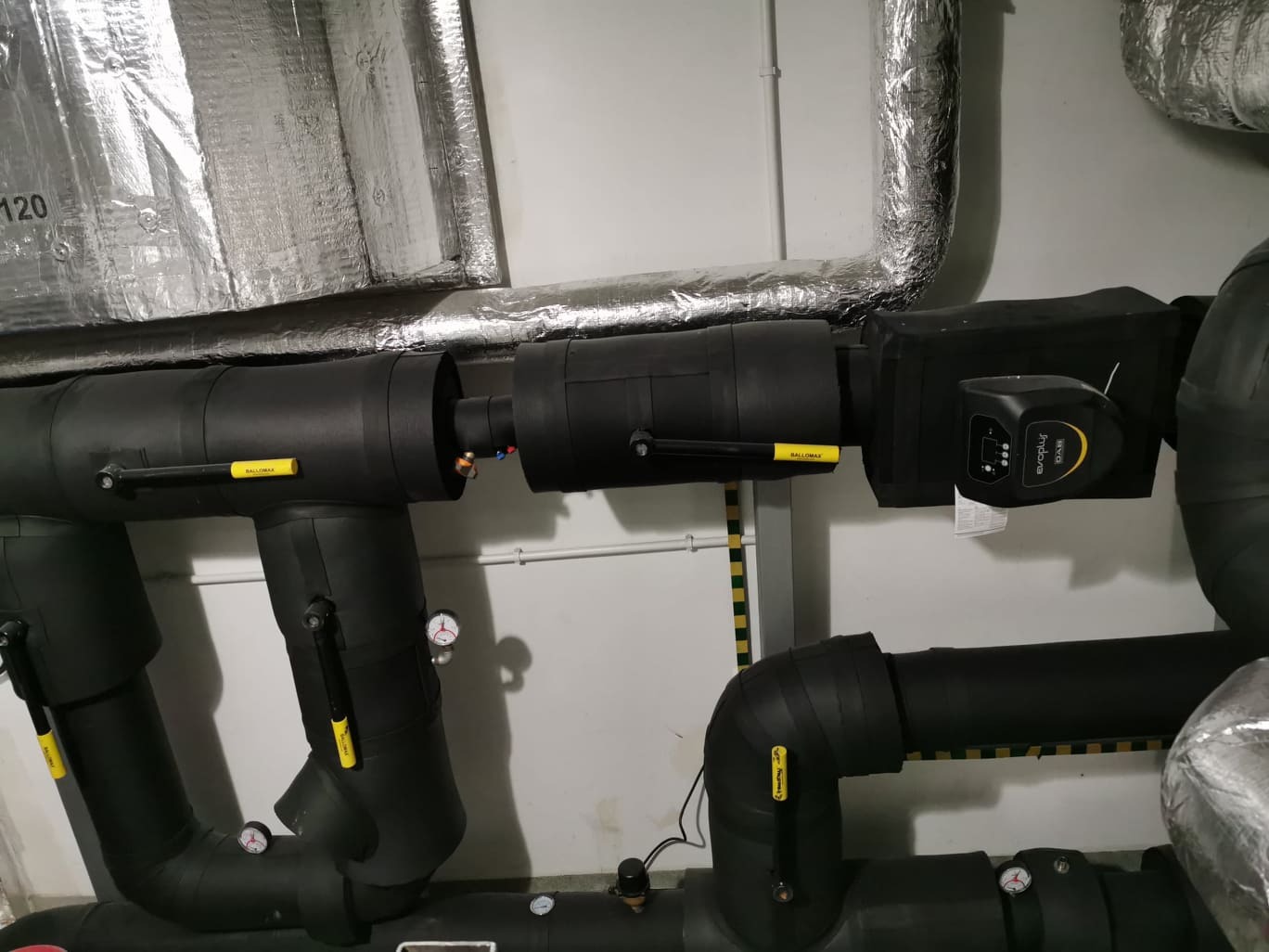 Izolowane rury grzewcze w piwnicy, widoczne zawory Ballomax, manometry oraz elektroniczna pompa obiegowa Evoplus Small z wyświetlaczem.