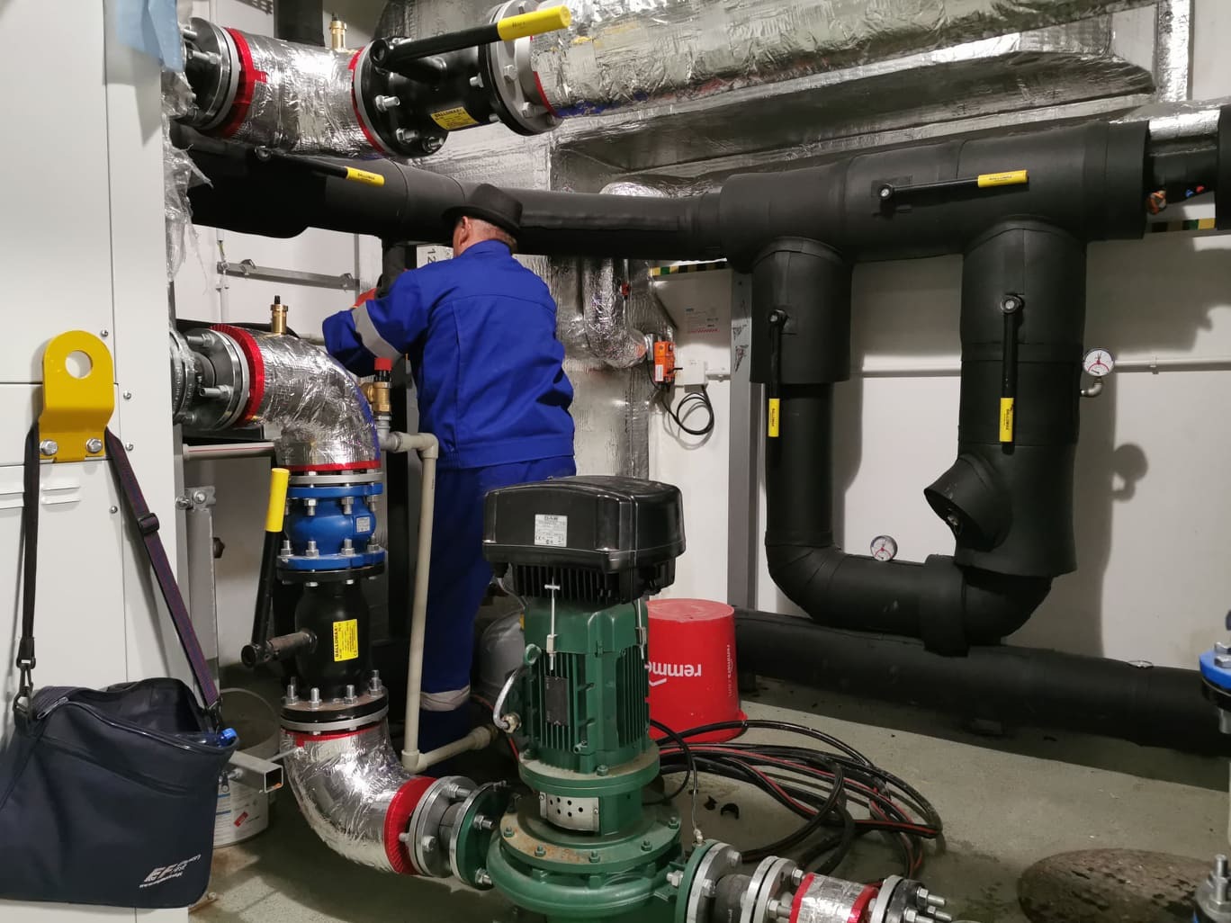 Pracownik w niebieskim kombinezonie i czarnym kapeluszu, w trakcie montażu rur w kotłowni, widoczne izolowane rury, zawory, pompy obiegowe i inne elementy instalacji grzewczej.
