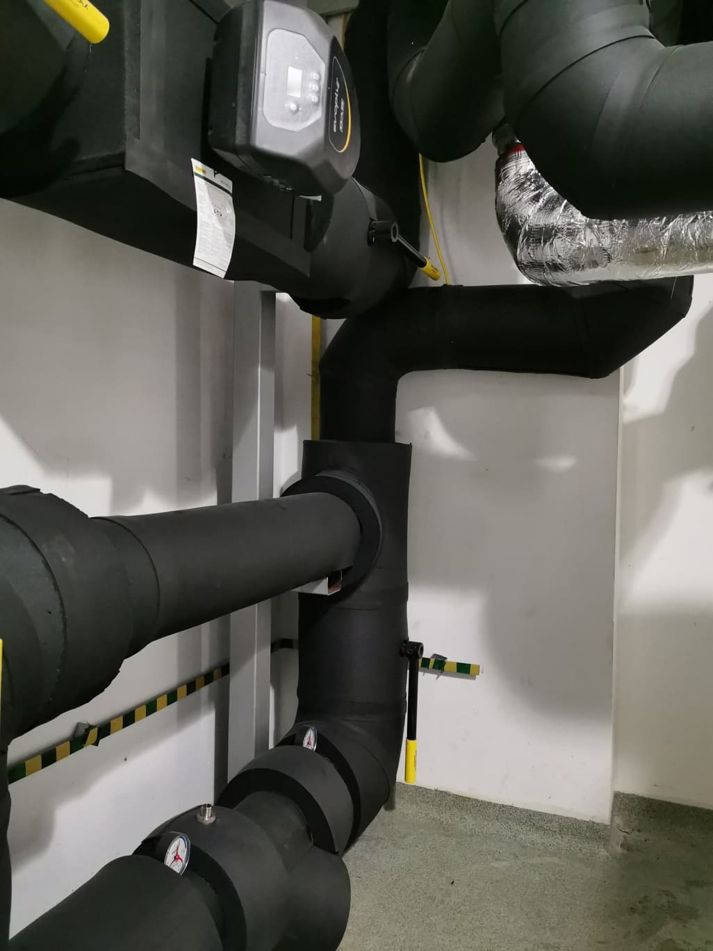 Izolowane czarne rury grzewcze z manometrami i zaworami, zamontowane w kotłowni z szarymi ścianami i podłogą, widoczny sterownik pompy obiegowej.