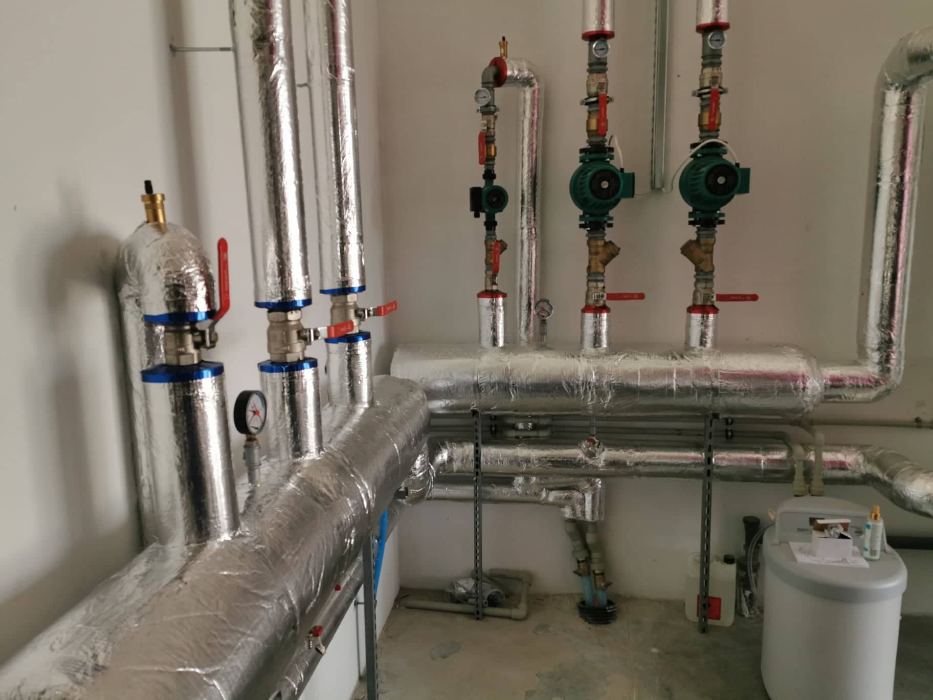 Izolowane rury grzewcze z zaworami i manometrami, widoczne pompy obiegowe, instalacja w jasnym pomieszczeniu technicznym, częściowo widoczny zmiękczacz wody.