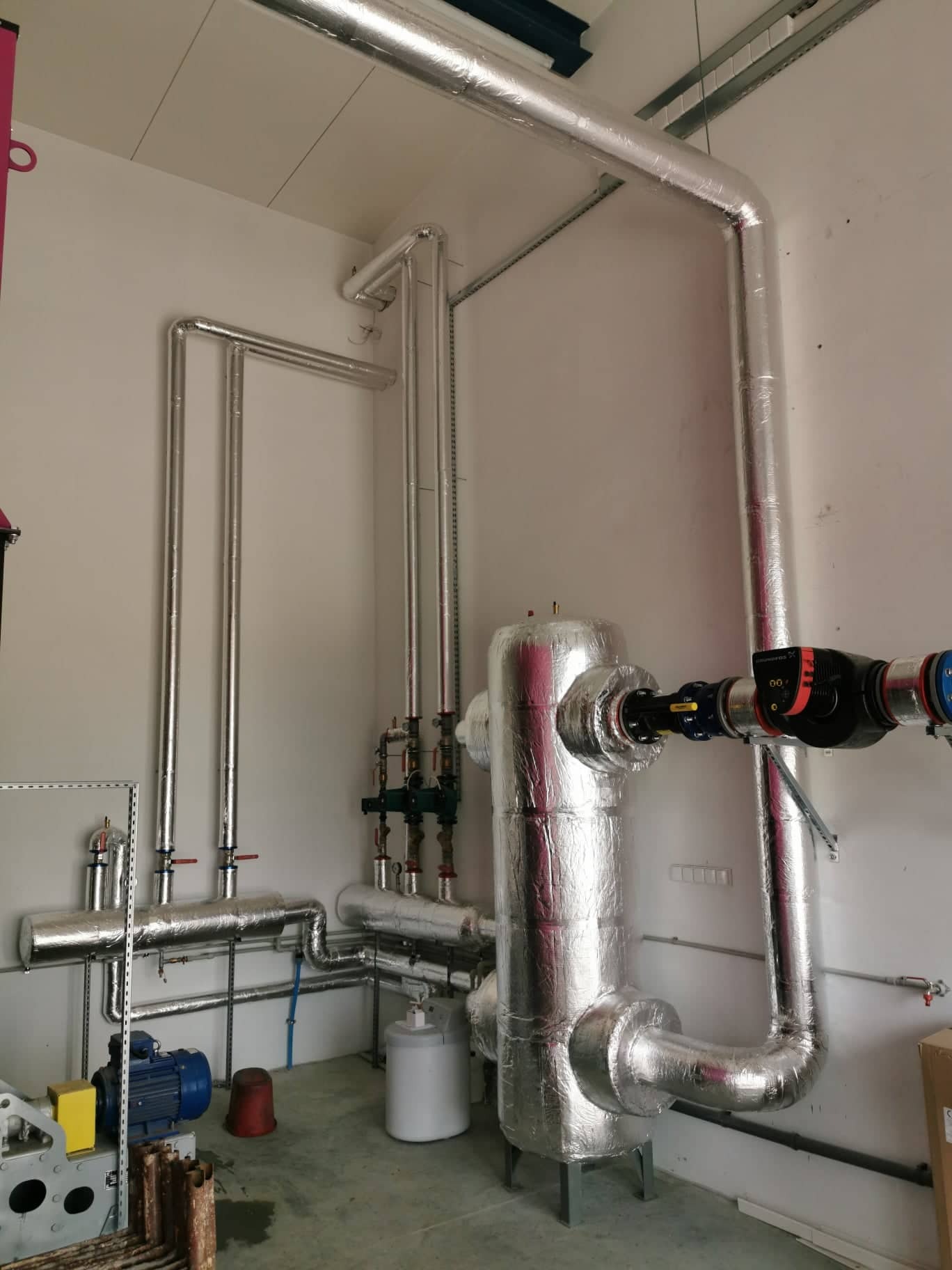 Kompleksowa instalacja grzewcza z izolacją termiczną, widoczne rury, zawory, zbiornik oraz silnik pompy obiegowej w pomieszczeniu technicznym.