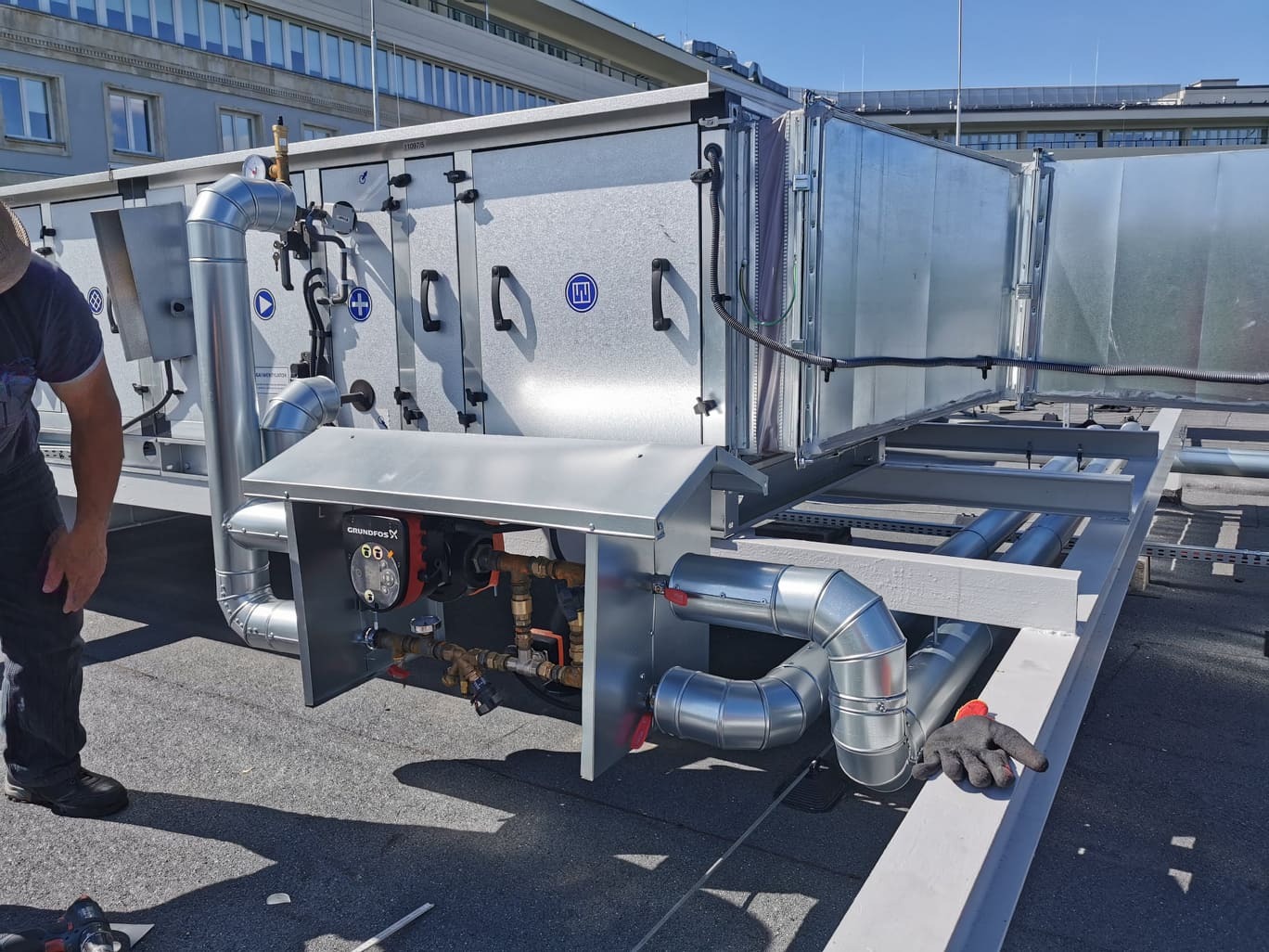 Montaż centrali wentylacyjnej na dachu budynku, widoczne kanały wentylacyjne, pompa obiegowa Grundfos i elementy instalacji hydraulicznej.