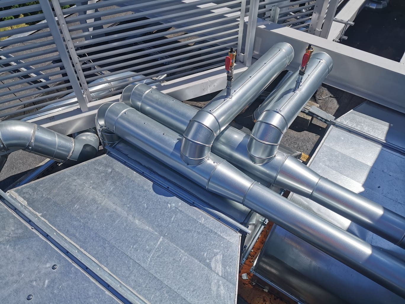 Błyszczące, metalowe rury wentylacyjne z zaworami na dachu, widziane z góry, z elementami konstrukcji wsporczej w tle.