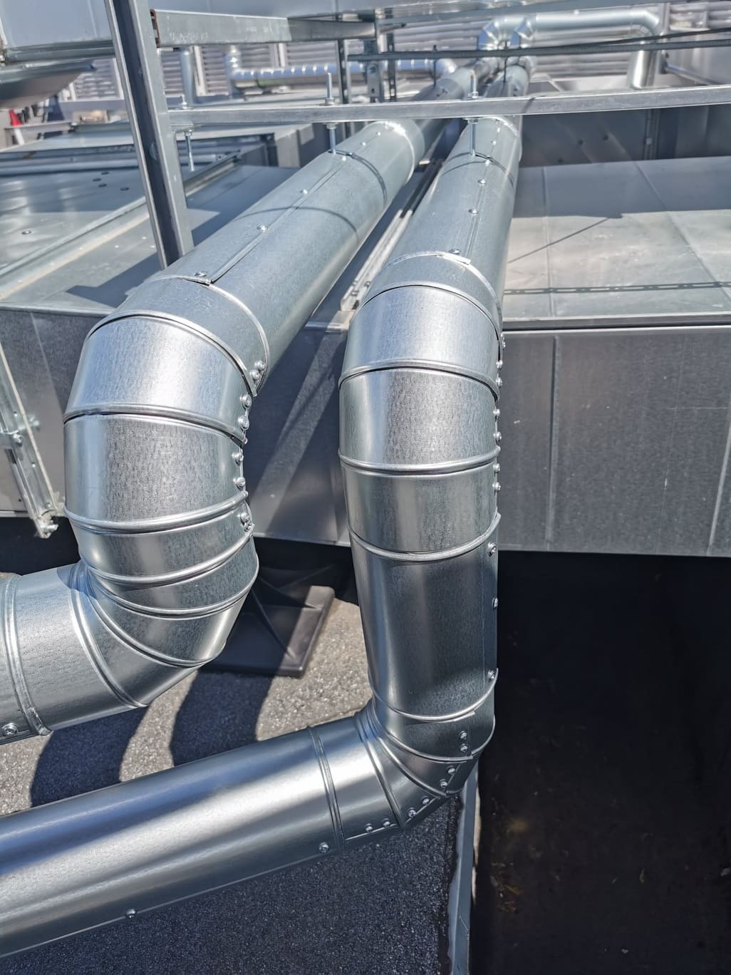 Dwa równoległe, ocynkowane przewody wentylacyjne z licznymi łączeniami nitowanymi, zamontowane na dachu pokrytym papą, widoczne elementy konstrukcji wsporczej.