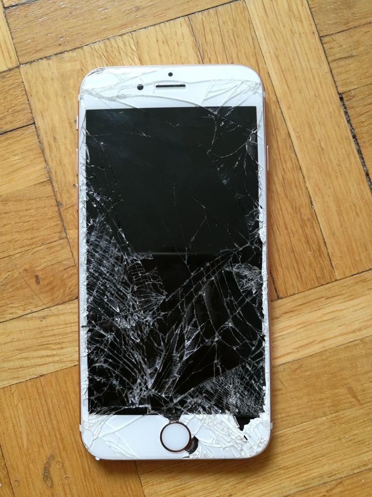 Smartfon z mocno potłuczonym ekranem leżący na drewnianej podłodze. Pęknięcia rozchodzą się promieniście od punktu uderzenia w dolnej części ekranu.