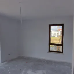 Surowe wnętrze pokoju z gładkimi, białymi ścianami, widokiem na plac budowy przez duże okno i wystającymi kablami z sufitu.